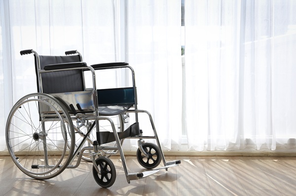 Empty wheelchair positioned in front of a sheer white curtain in a brightly lit room with wood flooring.