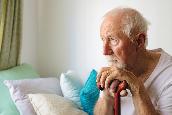 Elderly man sitting on a bed holding a cane, looking out with a serious expression in a softly lit room with pillows behind him.