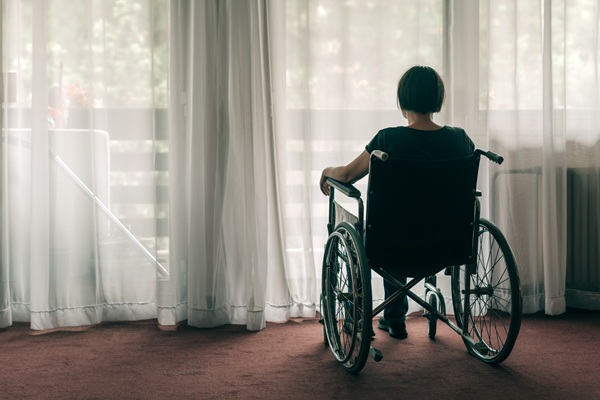 A female nursing home resident left alone in a wheelchair while facing a window with sheer curtains inside a nursing home.