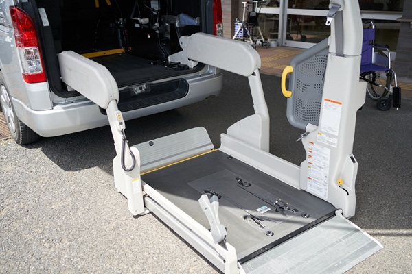 An open silver van features a mechanical wheelchair lift deployed to the ground, positioned outside a nursing facility.