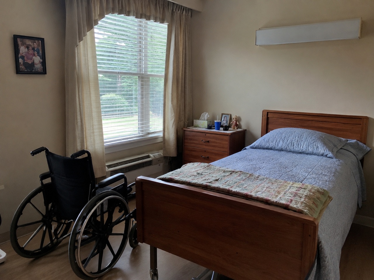 An empty Alabama nursing home room with an unmade bed, a wheelchair beside the window, personal items on the nightstand, and a family photo on the wall, depicting the aftermath of a nursing home resident's death and the potential for a wrongful death or neglect lawsuit against the facility.
